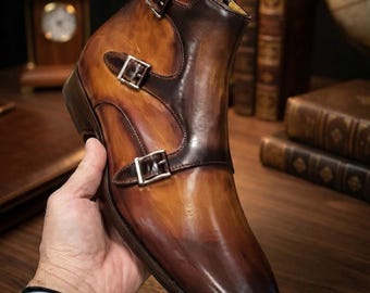 May include: A high-top boot in a burnished tan and dark brown leather finish, featuring three silver-toned metal buckles along the side, a pointed toe, a stacked heel, and a pull tab at the top of the ankle collar for ease of access.