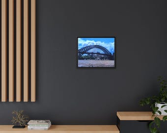 May include: Framed print of a large steel arch bridge spanning a body of water under a blue sky with clouds. The bridge is dark gray and has a curved design. The print is in a black frame.
