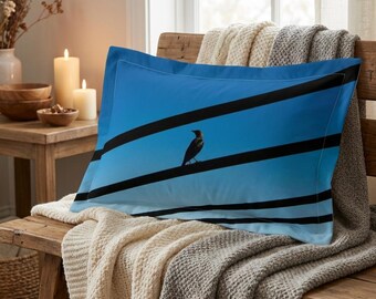 May include: A blue pillowcase featuring a bird perched on black lines against a bright blue sky. The pillow is resting on a wooden bench covered with a cream-colored knitted blanket and a gray blanket.