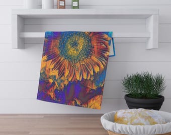 May include: A colorful kitchen towel featuring a vibrant sunflower design. The towel showcases a large, detailed sunflower in shades of yellow, orange, and blue. The towel is hanging on a white shelf above a wooden countertop.