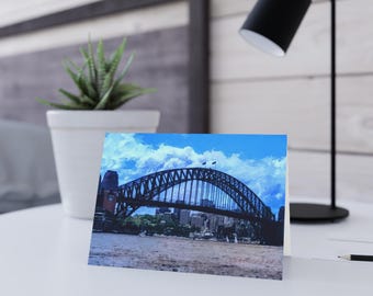 May include: A folded greeting card featuring a printed image of the Sydney Harbour Bridge spanning across a body of water under a bright blue sky with white clouds. The bridge is dark in color and shows architectural steel details.