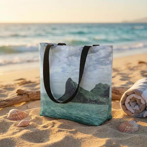 May include: A beach tote bag featuring a scenic landscape of mountains and ocean. The bag has black handles and is resting on a sandy beach with seashells and a rolled-up towel. The image on the bag depicts a coastal view.