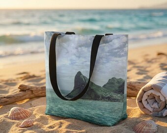 Puede incluir: Un bolso de playa con un paisaje de montañas y océano. El bolso tiene asas negras y está sobre una playa de arena con conchas marinas y una toalla enrollada. La imagen en el bolso muestra una vista costera.