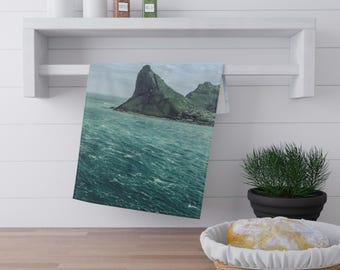 May include: A teal and green kitchen towel with a scenic landscape of a mountain and ocean. The towel is hanging on a white shelf with jars and spices. A loaf of bread sits in a basket on the counter.