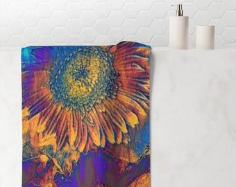 May include: A colorful bath towel featuring a large, stylized sunflower. The sunflower has yellow, orange, and blue hues, with a dark purple and blue background. The towel is displayed on a white shelf with a soap dispenser and a small cup.