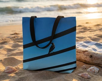 May include: Blue tote bag with black straps and a gradient design of blue stripes. A black bird silhouette is perched on a black stripe. The bag is on a sandy beach with seashells and driftwood.