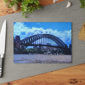 May include: A rectangular glass cutting board featuring a high-resolution print of the Sydney Harbour Bridge. The image displays the dark steel arch bridge spanning across the water with a bright blue sky and white clouds in the background.