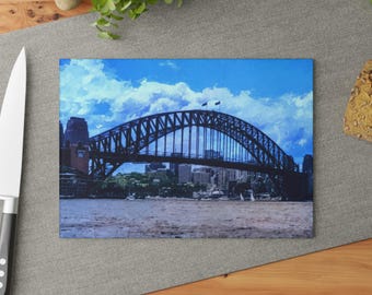 Puede incluir: Una tabla de cortar rectangular de vidrio con una impresión de alta resolución del puente Sydney Harbour Bridge. La imagen muestra el puente de arco de acero oscuro sobre el agua con un cielo azul brillante y nubes blancas de fondo.