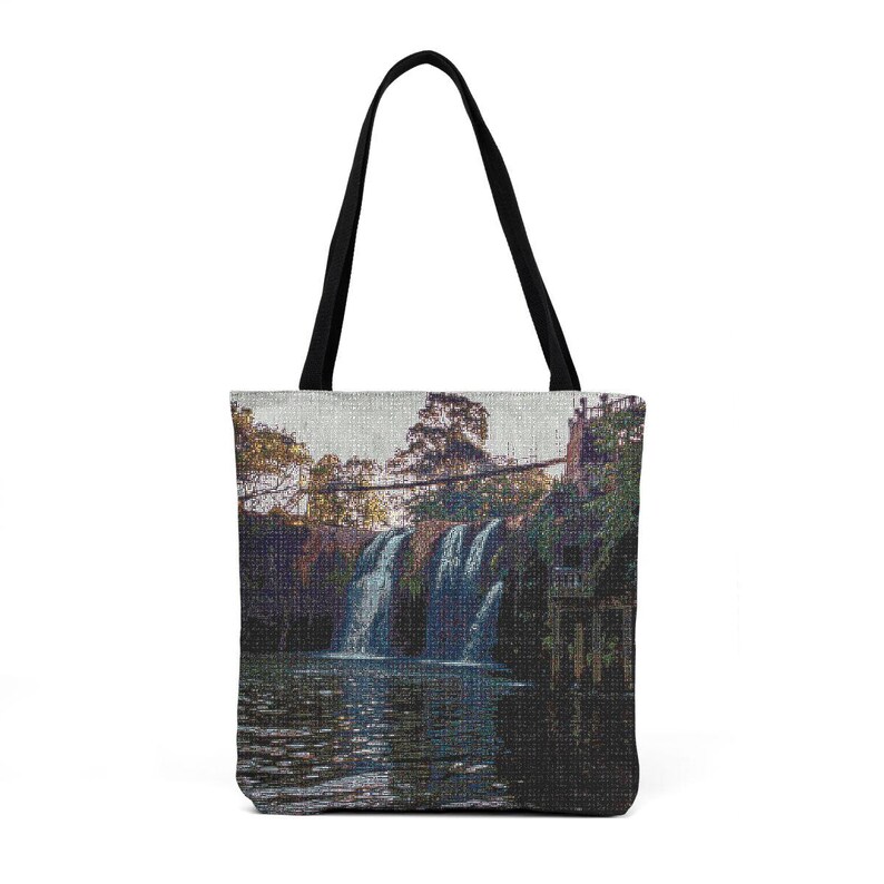 May include: Tote bag featuring a scenic waterfall landscape. The image shows a cascading waterfall flowing into a calm body of water, with lush greenery and a bridge in the background. The bag has black handles.