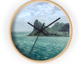 May include: A round wall clock with a light wood frame featuring a photographic print of a rocky mountain peak rising from a turbulent teal ocean under a cloudy sky. The clock face has three thin black hands indicating the time against the landscape.