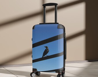 May include: A hard-shell rolling suitcase featuring a light blue sky background with two thick, horizontal black stripes. A small black bird is perched on the lower black stripe. The luggage has a black retractable handle and four wheels.