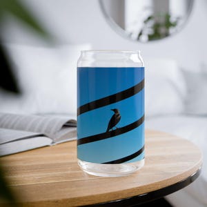 May include: A clear glass can-shaped drinking glass with a blue and black design. The design features a black bird perched on a black line against a blue gradient background. The glass sits on a wooden table.