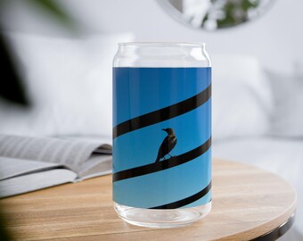 Puede incluir: Un vaso transparente con forma de lata con un diseño azul y negro. El diseño presenta un pájaro negro posado en una línea negra sobre un fondo degradado azul. El vaso está sobre una mesa de madera.