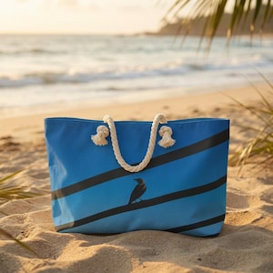 May include: A blue tote bag featuring black diagonal stripes and a bird illustration. It has a rope handle and is placed on a sandy beach. The background shows the sea and a palm frond.