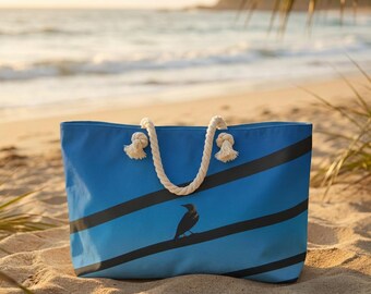 May include: A blue tote bag with black diagonal stripes and a bird graphic. The bag has a rope handle and is sitting on a sandy beach. The background shows the ocean and a palm frond.