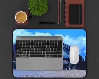 May include: A rectangular desk mat featuring a printed image of the Sydney Harbour Bridge under a bright blue sky with clouds. The mat is positioned beneath a silver laptop and a white computer mouse, creating a functional workspace setup.