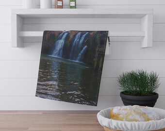 May include: A rectangular towel featuring a scenic waterfall image, hanging on a white shelf. The waterfall cascades into a dark body of water. A small green plant in a black pot and a basket with bread are in the foreground.