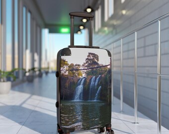 May include: A black rolling suitcase with a scenic waterfall image on the front. The suitcase has a retractable handle, four wheels, and black corner protectors. The waterfall image features a body of water and lush green trees.