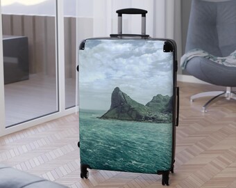 May include: A hard-shell rolling suitcase featuring a photographic print of a rocky mountain peak rising from a deep teal ocean under a cloudy sky. The luggage has a black frame, a retractable top handle, and four black spinner wheels at the base.
