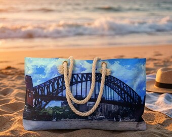 May include: A rectangular tote bag with a scenic image of a bridge over a city skyline, with a blue sky and clouds. The bag has rope handles and is resting on a sandy beach. A straw hat is on the sand.