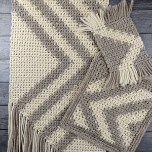 Op de afbeelding: Drie gehaakte tapijten met een chevron-patroon in beige en grijs. De tapijten hebben franjes aan de randen.