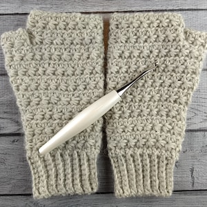 Op de afbeelding: Een paar beige gebreide vingerloze handschoenen met een witte haaknaald. De handschoenen hebben een textuurpatroon.