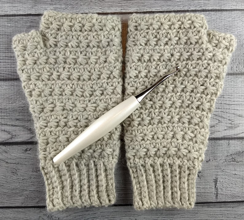 Op de afbeelding: Een paar beige gebreide vingerloze handschoenen met een witte haaknaald. De handschoenen hebben een textuurpatroon.