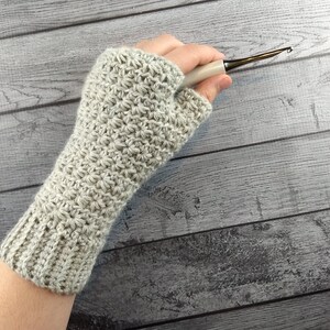 Op de afbeelding: Een grijze gehaakte vingerloze handschoen met een textuurpatroon. De handschoen wordt getoond op een hand die een haaknaald vasthoudt.