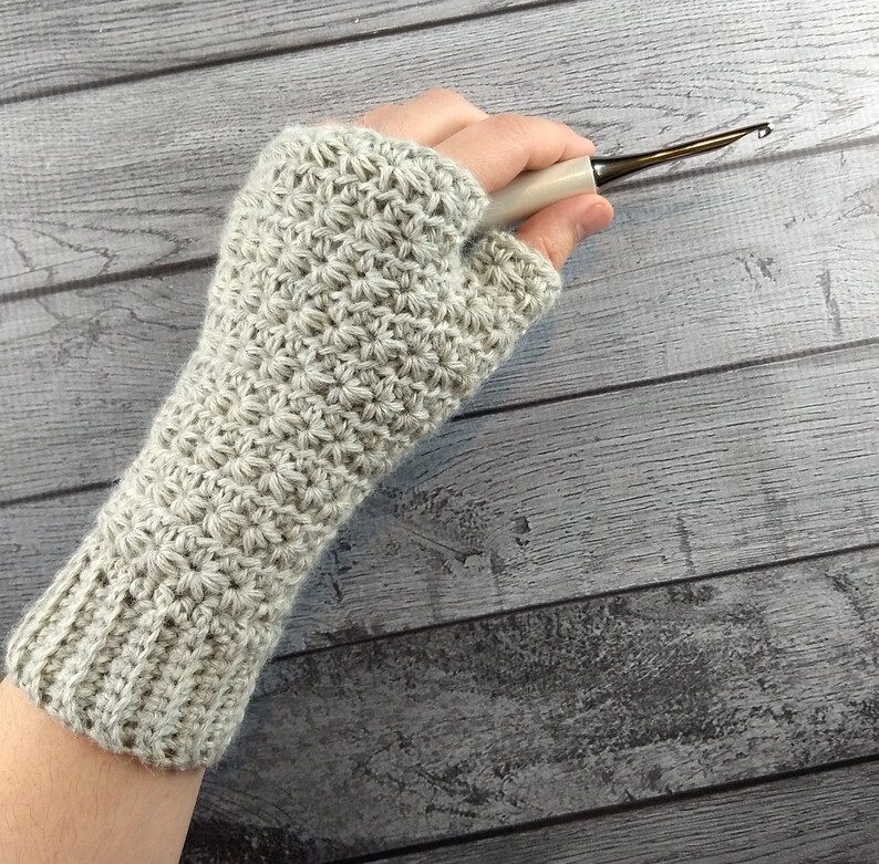 Op de afbeelding: Een grijze gehaakte vingerloze handschoen met een textuurpatroon. De handschoen wordt getoond op een hand die een haaknaald vasthoudt.