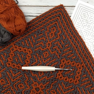 Könnte beinhalten: Nahaufnahme eines gehäkelten Quadrats mit einem geometrischen Muster in Orange und Grau. Das Quadrat ist mit einer Häkelnadel und Garn hergestellt.