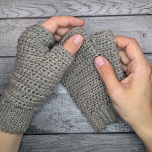 Puede incluir: Un par de guantes sin dedos de ganchillo grises. Los guantes están hechos de un hilo texturizado y tienen un puño acanalado. El pulgar está abierto y los guantes cubren la mano hasta los nudillos. Los guantes se muestran sobre un fondo de madera gris.
