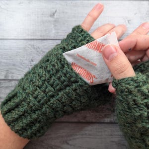 Könnte beinhalten: Grüne fingerlose Handschuhe aus Häkelarbeit mit einem Handwärmer-Paket im Inneren. Das Paket ist weiß mit orangefarbenem Text, der "HotHands.com" lautet.