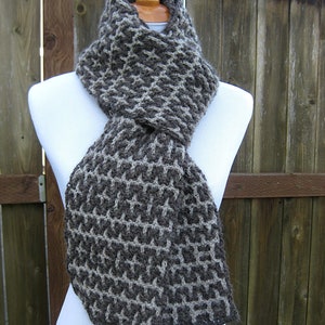 May include: A brown and white knitted scarf with a geometric pattern. The scarf is displayed on a white mannequin against a wooden background.