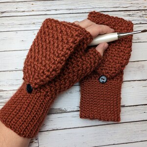 May include: A pair of brown crocheted fingerless gloves with a button on each cuff. The gloves are made with a textured stitch and are shown on a wooden background.