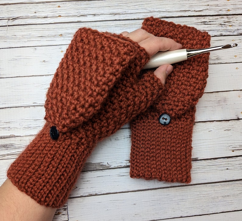 May include: A pair of brown crocheted fingerless gloves with a button on each cuff. The gloves are made with a textured stitch and are shown on a wooden background.