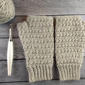 Op de afbeelding: Een paar beige gebreide vingerloze handschoenen met een textuurpatroon. De handschoenen liggen op een houten oppervlak met een bol garen en een haaknaald.