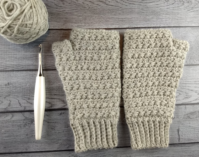 Op de afbeelding: Een paar beige gebreide vingerloze handschoenen met een textuurpatroon. De handschoenen liggen op een houten oppervlak met een bol garen en een haaknaald.