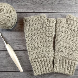 Op de afbeelding: Een paar beige gebreide vingerloze handschoenen met een textuurpatroon. De handschoenen liggen plat op een houten oppervlak met een bol garen en een haaknaald.