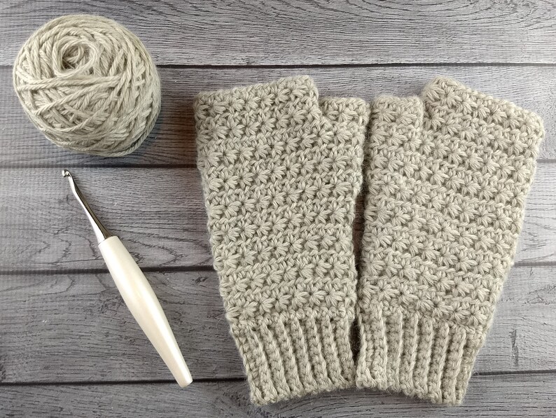 Op de afbeelding: Een paar beige gebreide vingerloze handschoenen met een textuurpatroon. De handschoenen liggen plat op een houten oppervlak met een bol garen en een haaknaald.