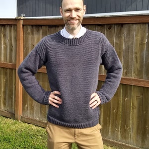 May include: A dark grey knitted jumper with a round neckline. The jumper is made with a textured stitch pattern.