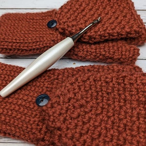 May include: A pair of rust-colored crocheted fingerless gloves with black buttons and a white crochet hook.