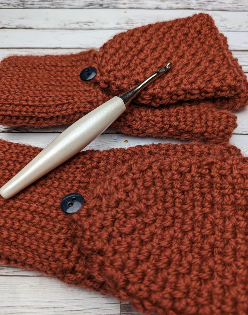 May include: A pair of rust-colored crocheted fingerless gloves with black buttons and a white crochet hook.