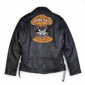 Puede incluir: Una chaqueta de cuero negra para motocicleta con un gran parche bordado en la espalda. El parche muestra una cara de gato estilizada dentro de una nube en forma de hongo con el texto ATOM CATS. La chaqueta incluye cordones laterales y cremalleras.