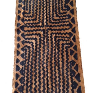 Puede incluir: Un camino de mesa textil rectangular con un patrón repetitivo de líneas verticales onduladas y formas triangulares en azul marino oscuro sobre un fondo marrón claro natural. La tela tiene bordes crudos sin rematar en los extremos.