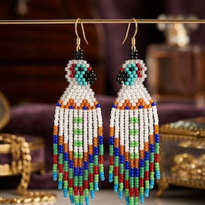 Puede incluir: Un par de pendientes de cuentas con un diseño colorido. Los pendientes presentan cuentas blancas, azules, verdes, rojas y naranjas. La parte superior tiene un diseño negro, rojo y turquesa. Los pendientes cuelgan de ganchos dorados.