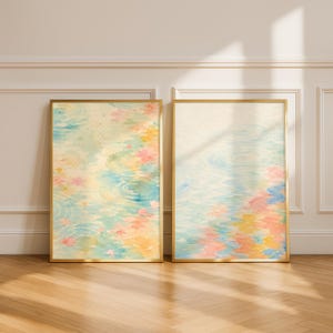 以下が含まれることがあります： 金色のフレームに入った2つの水彩画。アートワークは、ピンク、黄色、青などのパステルカラーの水面のさざ波と花の要素の柔らかく抽象的なデザインを特徴としています。絵画は白い壁と木製の床に展示されています。