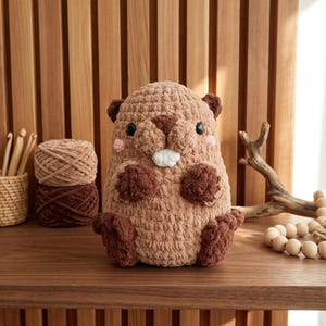 以下が含まれることがあります： 茶色の糸で手編みされたビーバーのぬいぐるみ。小さな黒い目、白い歯、ピンクの頬が特徴です。ビーバーは木製の棚の上に置かれており、背景には毛糸と編み棒が見えます。