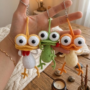 Puede incluir: Tres llaveros de rana hechos a mano con ganchillo en amarillo, verde y naranja. Cada rana tiene ojos grandes y saltones y patas pequeñas. La rana amarilla tiene una pajarita roja. Sobre una superficie de madera.