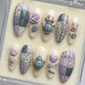 May include: A set of ten press-on nails featuring a cream base with various designs including patchwork patterns in purple, green, and white, a 3D purple rose, a 3D teapot, a 3D heart, a seed packet illustration, and the text BLOOM.