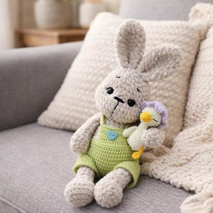 May include: A handmade crochet bunny doll in light gray, wearing green overalls with a blue heart-shaped button. The bunny holds a small duck doll with a purple hat. The dolls are sitting on a gray couch with a white pillow and blanket.